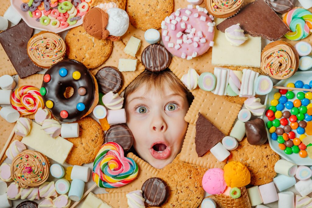 Funny 5 years old girl lying under plenty of sweet foods. Kids face surrounded by unhealthy food. To